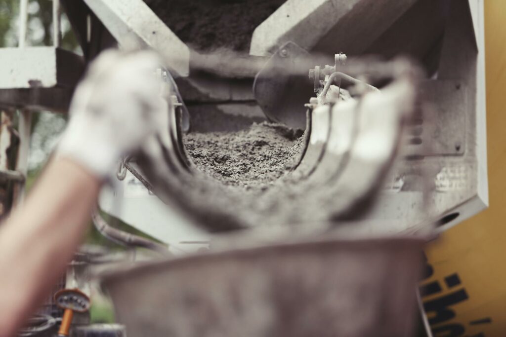 This picture shows the scene of a construction site. In the foreground a hand wearing work gloves is carrying a bucket. In the background a cement truck is visible. Cement is flowing from the truck into the bucket. budowie