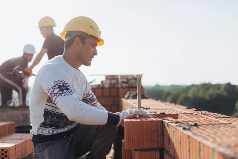 Construction worker laying bricks outdoors in sunny weather with lush greenery in background.